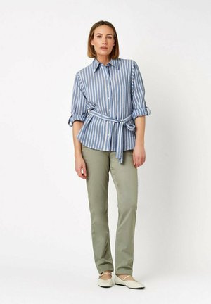 Donna in piedi che indossa una camicia a righe blu e bianca con cintura, pantaloni verde oliva e scarpe basse beige su sfondo bianco.