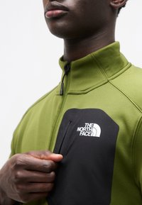 Πράσινο φλις μπουφάν με ψηλό γιακά, μαύρη τσέπη στο στήθος με φερμουάρ και λευκό λογότυπο. Το ύφασμα φαίνεται υφασμάτινο και απαλό.