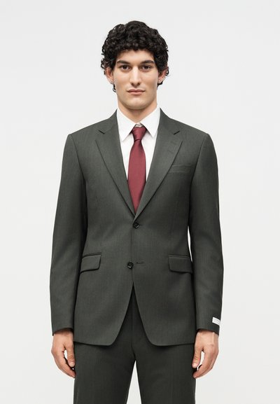 Νέος άνδρας με σγουρά μαλλιά, φορώντας σκούρο γκρι κοστούμι, λευκό πουκάμισο και μπορντώ γραβάτα, στέκεται απέναντι σε ένα απλό φωτεινό φόντο.