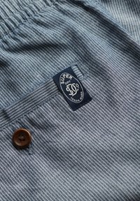 Blauw gestreepte stof met een textuurpatroon. Bevat een bruine knop en een marineblauwe label met het "Superdry"-logo. Het ontwerp is casual en eenvoudig.