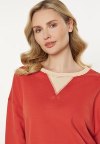 Punainen huppari, jossa on beige kaulus ja V-muotoinen pääntie, pehmeä kangas ja näkyvät ompelu yksityiskohdat. Mallin kasvot osittain näkyvissä.