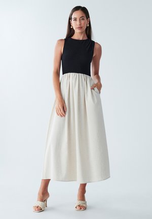 Maxime dress, jossa on tiukka musta smokkiliiviosa ja virtaava kermaisa hame. Sisältää taskut; mallilla on päällään beige korokepohjainen sandaalit.