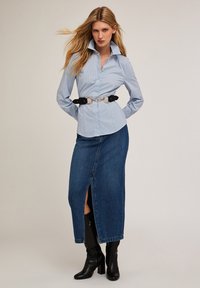 Camisa azul a rayas con cuello alto, combinada con una falda de mezclilla oscura con una abertura lateral, ceñida con un cinturón negro y herrajes dorados.