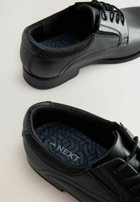 Chaussures en cuir noir avec une finition lisse, bout rond et fermeture à lacets. La semelle intérieure texturée en bleu marine présente un motif géométrique et le nom de la marque.