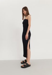 Melnas strapless maksi kleitas ar teksturētu audumu un augsto sānu šķēlumu, apvienotas ar melnām masīvām sandālēm ar divām sprādzēm.