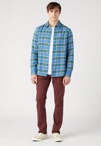 Camisa de cuadros azul y verde, sobre una camiseta blanca, combinada con pantalones burdeos y zapatillas blancas, de pie contra un fondo neutro.