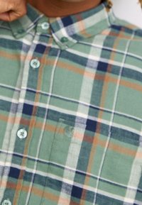 Grünes Karoshirt mit blauen und orangefarbenen Akzenten. Knopfverschluss, kurzer Kragen und eine Brusttasche auf der linken Seite. Weicher Stoff.