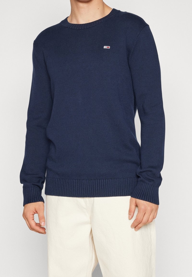 Homme portant un pull bleu marine à col rond avec un petit logo rectangulaire sur la poitrine, associé à un pantalon écru.