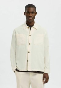 Giacca button-up beige chiaro con due tasche sul petto e colletto. Realizzata in tessuto texturizzato, presenta bottoni marroni e una vestibilità comoda.
