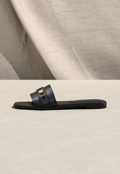 Μαύρο επίπεδο σανδάλι τύπου σλάιντ με δύο φαρδιές λωρίδες και χρυσό κυκλικό έμβλημα, τοποθετημένο πάνω σε φόντο από μπεζ ύφασμα και επιφάνεια.