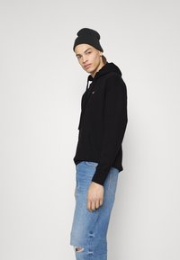 Schwarzer Kapuzenpullover mit Tasche, verfügt über gerippte Bündchen, getragen mit hellblauen, distressed Jeans und einer dunklen Mütze, steht vor einem hellen Hintergrund.