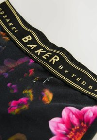 Tela negra con patrones florales en rosa y amarillo. La cinturilla elástica presenta un texto dorado: "BAKER by Ted Baker." Textura suave.