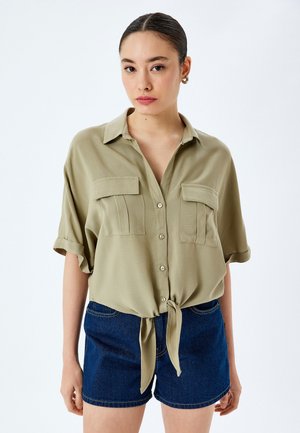 Camicia verde oliva a maniche corte con chiusura a bottoni, due tasche sul petto e orlo annodato, abbinata a pantaloni shorts di denim blu scuro.