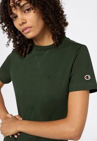 Młoda kobieta z kręconymi włosami, ubrana w ciemnozielony t-shirt Champion z naszywkami z logo na klatce piersiowej i rękawie, z rękami skrzyżowanymi na talii.