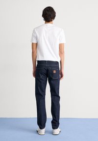 Dark blue denim jeans with a straight cut, featuring yellow stitching and two back pockets. Worn with a plain white t-shirt and light shoes.