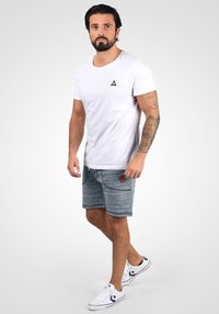 Camiseta blanca de cuello redondo con pequeño logotipo triangular negro; pantalones cortos azul jaspeado con bolsillos y detalles en cuero marrón; zapatillas blancas.
