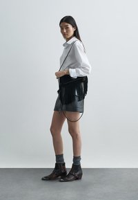 Melna frēze, šķērsgriezuma soma, balts apkakles krekls, pelēki šorti, pelēkas zeķes un brūni rakstaini potīšu zābaki. Minimālistisks fons.