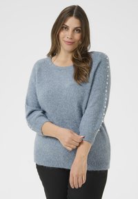 Naine kannab pehmet sinist sviitrit, millel on säravad nööbid hiha ääres, seistes ühetoonilise valge tausta ees ja naeratades õrnalt.