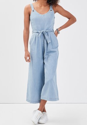 Combinaison en denim bleu clair avec des jambes larges, bretelles réglables, taille cintrée et ceinture à nouer. Présente des accents en métal argenté.