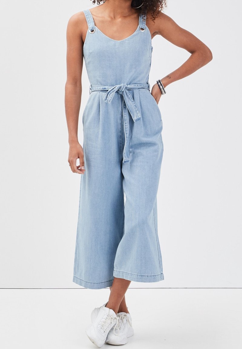 Light blue denim jumpsuit with wide legs, adjustable shoulder straps, a fitted waist, and a tied belt. Features silver hardware accents.