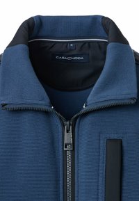 Blauwe ritsjack gemaakt van textuurstof met een hoge kraag, zwarte accenten, een zichtbare rits en een vierkante flap op de voorkant.