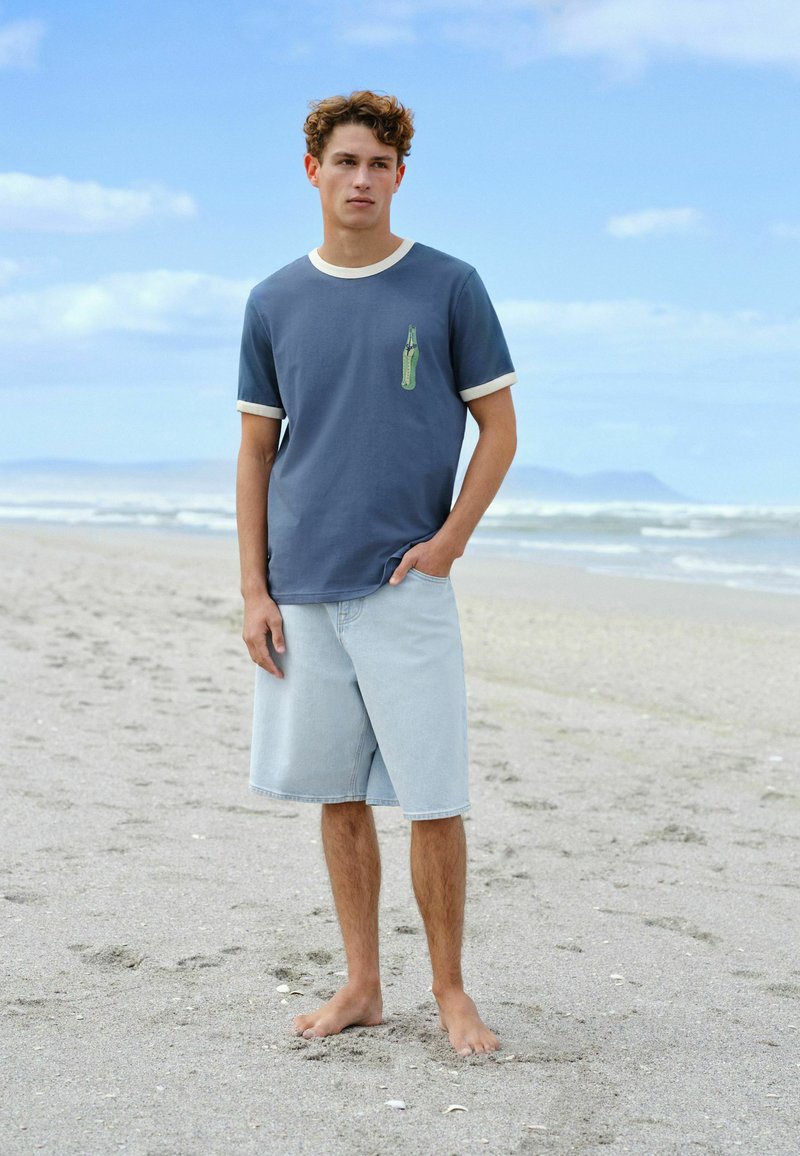 Jeune homme aux cheveux bouclés debout pieds nus sur une plage de sable, portant un t-shirt bleu avec un imprimé bouteille et un short en jean clair.