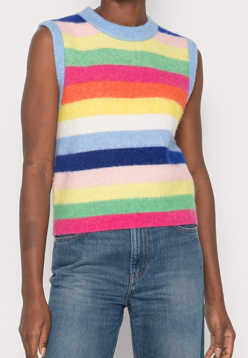 Pull sans manches en matière duveteuse avec des rayures horizontales arc-en-ciel porté avec un jean bleu sur une personne devant un fond uni.