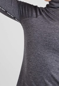 Haut à manches longues gris avec un col montant, fabriqué dans un matériau doux. Présente un logo noir "Nike Pro" le long de la manche et une coupe ajustée.