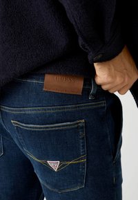 Ciemnoniebieskie dżinsy z żółtym przeszyciem, z brązową skórzaną łatą z logo z tyłu oraz czerwoną trójkątną metką na kieszeni.