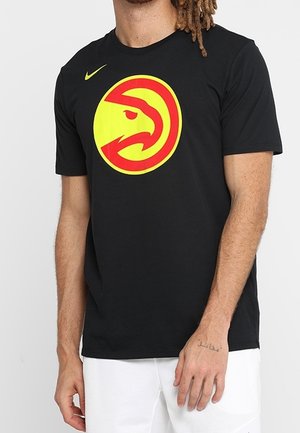 Mann trägt ein schwarzes T-Shirt mit großem gelbem und rotem Falkenlogo sowie kleinem gelbem Nike-Swoosh auf der Brust, kombiniert mit weißen Shorts.