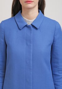 Blue textured coat with a collar, front buttons, and long sleeves. The fabric has a subtle pattern and a structured fit.