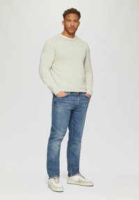 Hellgrauer Strickpullover mit Rundhalsausschnitt, kombiniert mit blauen Jeans und beigen Sneakers. Stehende Pose vor einem einfarbigen Hintergrund.