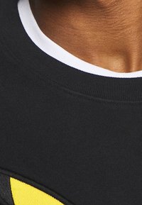 Felpa nera con un design giallo, caratterizzata da un collo rotondo bianco a contrasto. Il tessuto appare morbido e con una trama liscia.