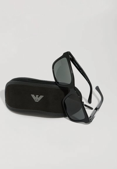 Ochelari de soare negri cu lentile închise și formă rectangulară, așezați pe un etui întunecat din gamă suede, având un logo argintiu și închidere cu fermoar.