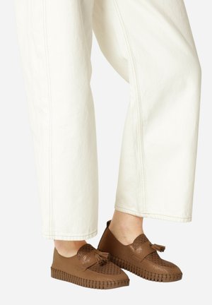 Zapatos marrones de fácil puesta con un empeine texturizado que presenta perforaciones y borlas decorativas, combinados con pantalones de pierna ancha blancos.