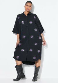 Zwarte button-up jurk met grote paarse oogmotieven en stralende zonnestraalpatronen. Heeft korte mouwen en een gebogen zoom. G gedragen met zwarte laarzen.