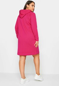 Fuksian väri hupparimekko pitkillä hihoilla, joka ulottuu reiden puoliväliin, ja siinä on suora leikkaus sekä pehmeä rakenne. Visuaalista ilmettä täydentävät valkoiset lenkkarit.