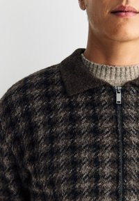 Veste zippée à carreaux marron et gris avec un col foncé, présentant un tissu texturé et un col côtelé en dessous.