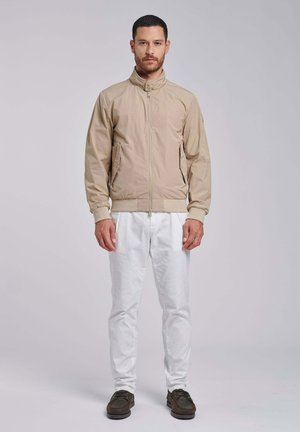 Man staat, draagt een beige jas met rits, witte broek en donkerbruine schoenen tegen een effen lichte achtergrond.