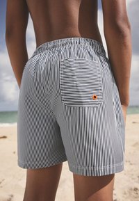 Tumesinise ja valge triibulised ujumispüksid, millel on üks tagatasku oranži aktsendiga. Elastne vöökoht, kergest kangast ning lõdva lõikega.