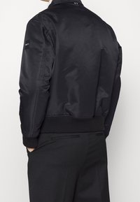 Svart bomberjacka i slät tyg, med ribbad krage och manschetter, en logotypapplikation på ärmen och en smal passform.