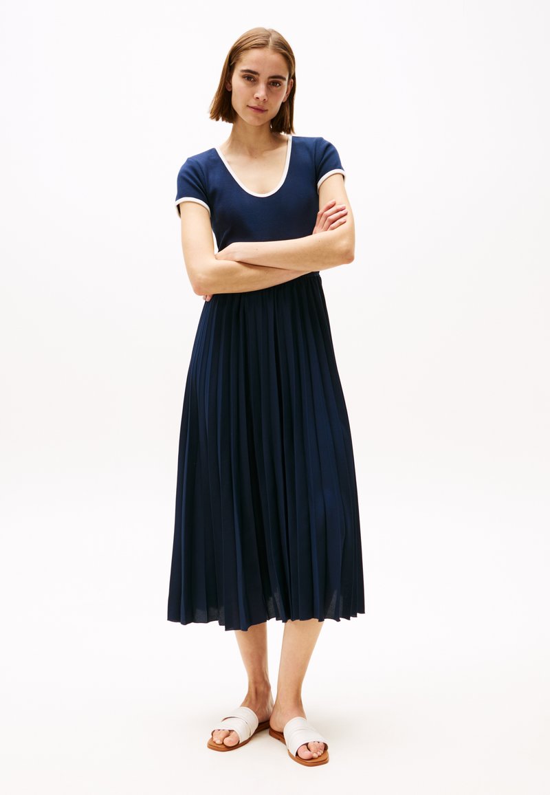 Robe plissée bleu marine avec un haut ajusté et un décolleté en V large, dotée de manches courtes avec des accents blancs. Le mannequin porte des sandales blanches à glissière.