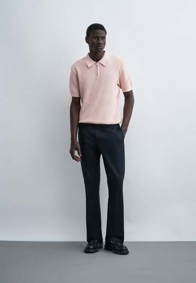 Άνδρας με σκουρόχρωμο δέρμα, που φοράει ένα ανοιχτό ροζ κοντομάνικο πλεκτό πόλο πουκάμισο, μαύρο παντελόνι και μαύρα μοκασίνια, στέκεται μπροστά από ένα απλό ανοιχτό φόντο.