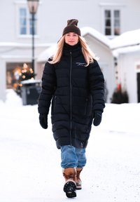 Czarna puchowa kurtka z zamkiem, logo na piersi, w połączeniu z jasnoniebieskimi dżinsami i brązowymi śniegowcami. Zimowa sceneria z widocznym śniegiem.