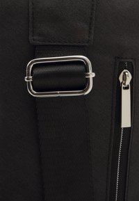 Dettaglio di una borsa di stoffa nera con una fibbia rettangolare argento, una tracolla texturizzata e una zip laterale con un tirante metallico.