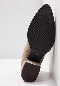 Bottine beige en daim à talon avec semelle noire nervurée, bout pointu, et taille 37 visible sur la semelle.