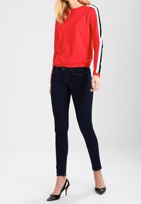 Haut à manches longues rouges avec des rayures latérales noires et blanches, associé à un jean skinny bleu foncé et des escarpins noirs. Tissu lisse et design ajusté.