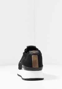 Black athletic shoe with a textured knit upper, gold stripes on the heel tab, and a white midsole. Features a rounded toe and flexible sole.