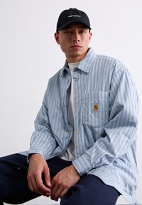 Chemise à rayures bleu clair en coton, dotée d'une poche poitrine avec un logo, accompagnée d'une casquette noire et d'un pantalon noir.