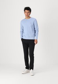 Lys blå bomullssweater med rund hals og lange ermer, kombinert med slanke svarte bukser og hvite joggesko. Diskret logo-detalj på brystet.
